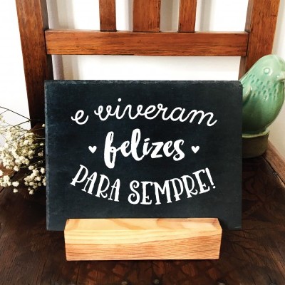 Placa decorativa com texto em branco sobre suporte de madeira, sobre cadeira de madeira com vaso e flores secas.