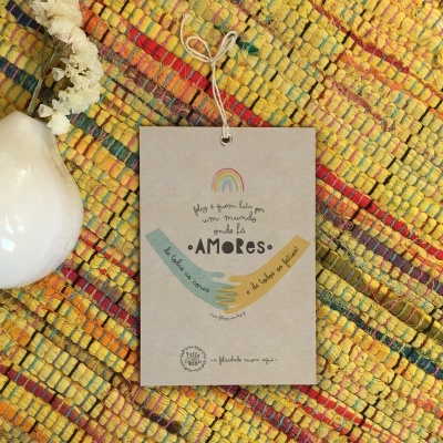 Etiqueta em papel com ilustrações e texto sobre tapete colorido, com vaso branco e flor seca ao lado.