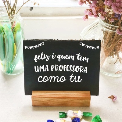 Cartão negro com mensagem em branco num suporte de madeira, flores em jarros de vidro e balas coloridas sobre mesa branca.