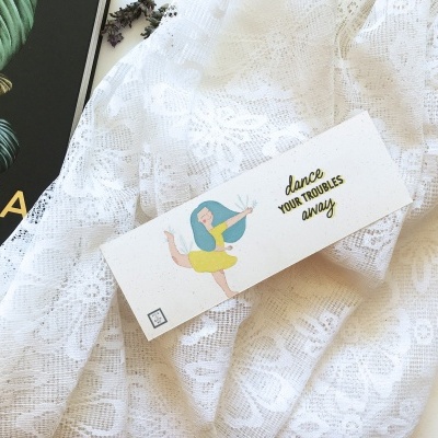Cartão com ilustração de menina a dançar e frase inspiradora sobre tecido rendado branco e revista parcial.