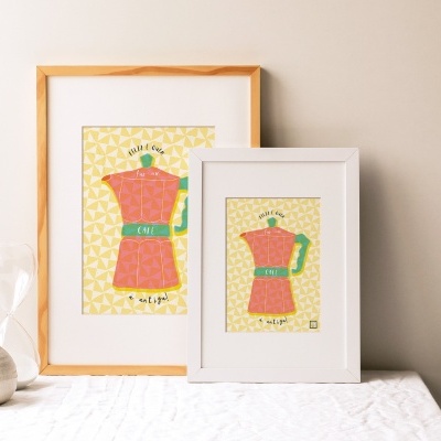 Dois quadros com ilustrações de cafeteiras vermelhas em molduras de madeira clara e branca sobre mesa com vaso e ampulheta.