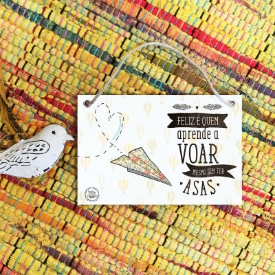 Placa decorativa branca com texto em português e avião de papel colorido sobre uma manta ou tapete colorido