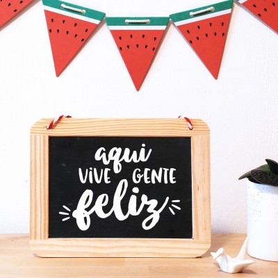 Quadro negro com frase feliz, vaso branco com planta e guirlanda de melancia na parede