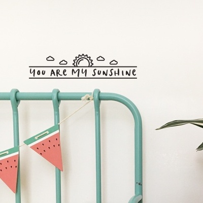 Cabeceira de cama verde água com decoração de melancia, autocolante de parede 'YOU ARE MY SUNSHINE' e planta ao lado.