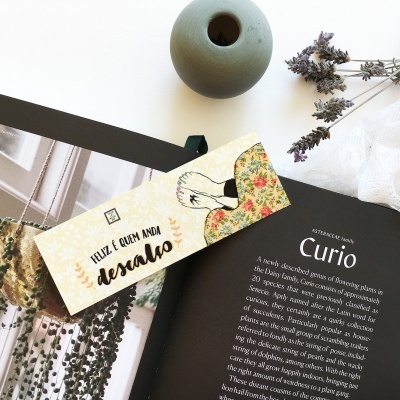 Livro aberto com texto, cartão com frase em português, vaso cinza, flores secas e tecido branco