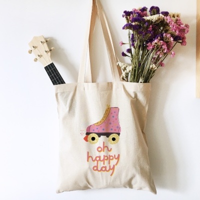 Saco de tecido cru com desenho de patim e texto 'oh happy day', flores secas e guitarra numa parede branca