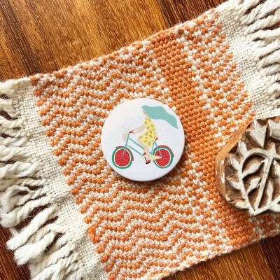 Botão redondo com ilustração de girafa numa bicicleta com guarda-chuva, sobre tecido laranja e branco, junto a carimbo de madeira