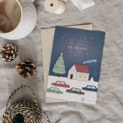Cartão de felicitações com ilustração de casa, árvores e carros sobre tecido cinza ao lado de cones de pinheiro, bola de fio e caneca