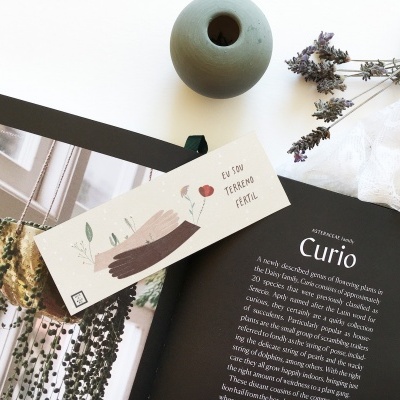 Revista aberta com texto 'Curio', marcador creme com folhas e balão vermelho, vaso cinza e ramos secos sobre pano branco