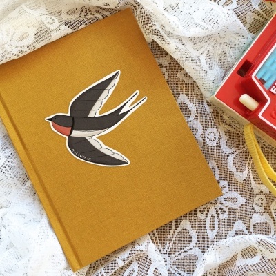 Livro de capa dura amarelo com desenho de andorinha e câmara Fisher-Price vermelha sobre renda branca