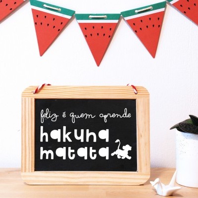 Quadro negro com texto inspiração hakuna matata numa mesa de madeira clara com decoração de melancia e planta em vaso branco