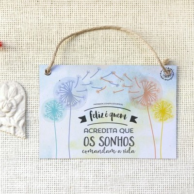 Placa decorativa de madeira com texto motivacional e desenho de dente-de-leão, pendurada ao lado de coração branco sobre tecido bege.