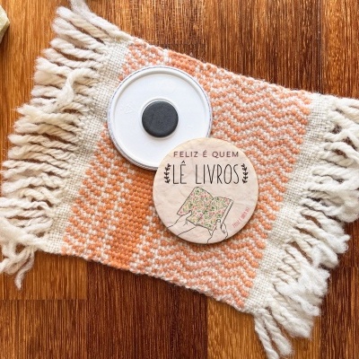 Marcador de livros redondo de madeira com texto FELIZ É QUEM LÊ LIVROS sobre tapete de algodão laranja e branco com franjas.