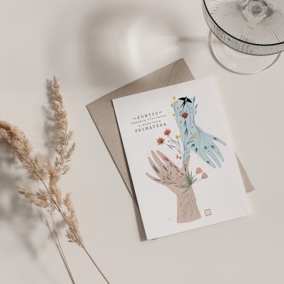 Cartão branco com ilustração de mãos e flores, envelope castanho, ramo seco e copo de vidro