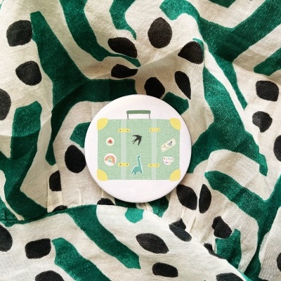 Botão redondo com desenho de mala verde sobre tecido branco com padrão verde e manchas negras