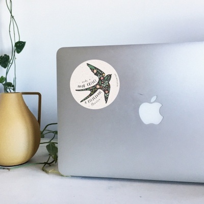 Portátil cinzento com autocolante de pássaro ao lado de vaso amarelo com planta verde