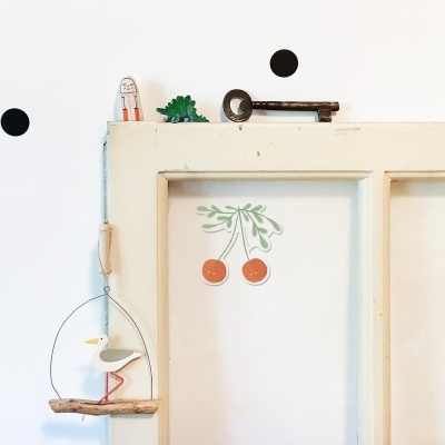 Janela antiga de madeira branca com autocolante de cerejas e móbile decorativo azul na parede branca com pontos pretos.