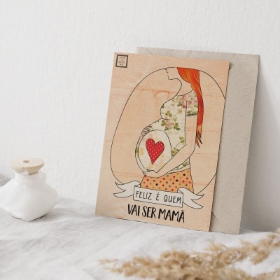 Cartão ilustrado de mulher grávida com texto 'FELIZ É QUEM VAI SER MAMÃ' em superfície branca com frasco decorativo.