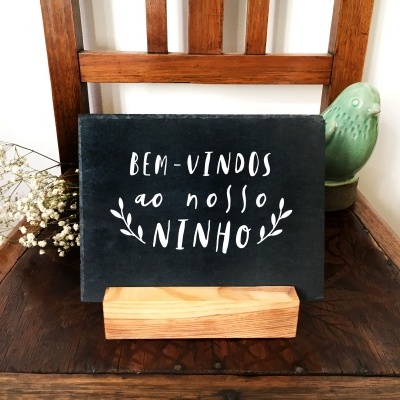 Placa decorativa preta com texto em branco sobre base de madeira numa cadeira de madeira escura com flores e ornamento de pássaro