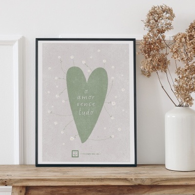 Pôster com coração verde e frase em moldura preta numa mesa de madeira com vaso branco e flores secas