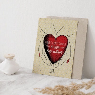 Cartão ilustrado com mãos e coração contendo texto, sobre superfície branca com vaso e planta seca.