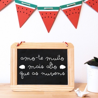 Lousa preta com moldura de madeira e texto em branco, vaso com planta e bandeirola decorativa de melancia