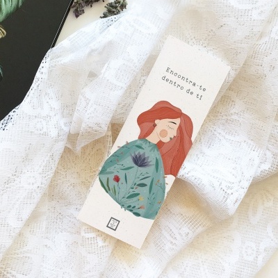 Livro pequeno ilustrado com mulher ruiva e flores sobre tecido branco rendado