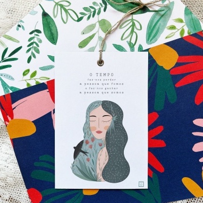 Três cartões decorativos coloridos com padrões florais e ilustração de mulher, amarrados com cordão sobre tecido rendado.