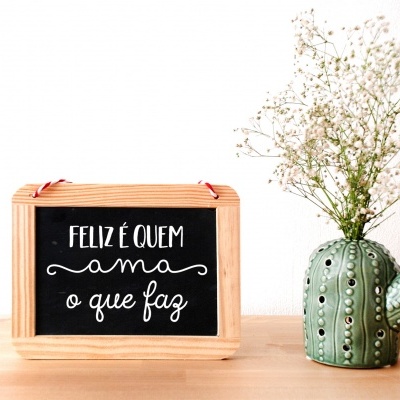 Placa de madeira com texto motivacional ao lado de vaso em forma de cacto com flores brancas