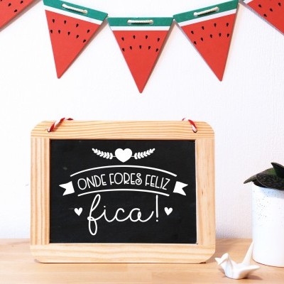 Placa de ardósia com moldura de madeira, texto em branco, vaso branco com planta e decoração de bandeirolas de melancia na parede