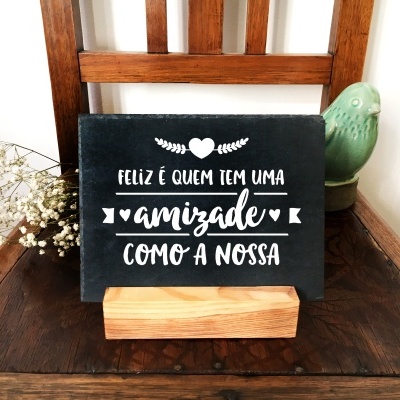 Placa preta com texto branco sobre suporte de madeira numa cadeira de madeira escura com vaso verde e flores secas.