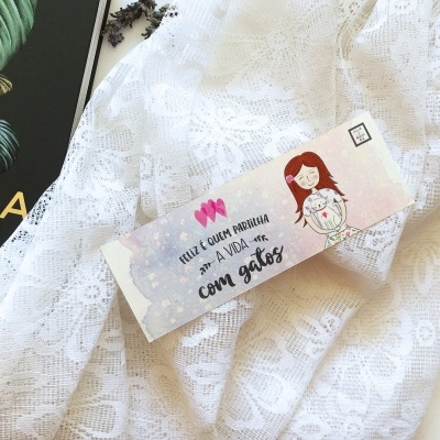 Marcador de páginas com ilustração de rapariga e texto, sobre tecido branco rendado e capa de livro preta