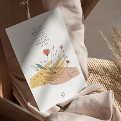 Cartão branco com ilustração de mãos e flores sobre tecido rosa claro numa cadeira de madeira