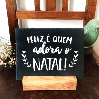 Placa decorativa preta com texto natalício em suporte de madeira sobre cadeira de madeira