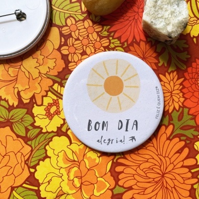 Dois botons redondos, sendo um com desenho de sol e texto 'BOM DIA', sobre toalha floral colorida