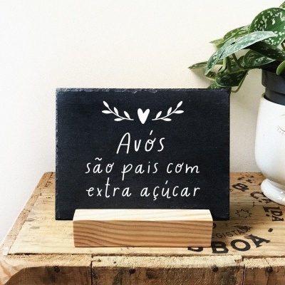 Placa decorativa preta com texto branco sobre suporte de madeira ao lado de vaso branco com planta verde