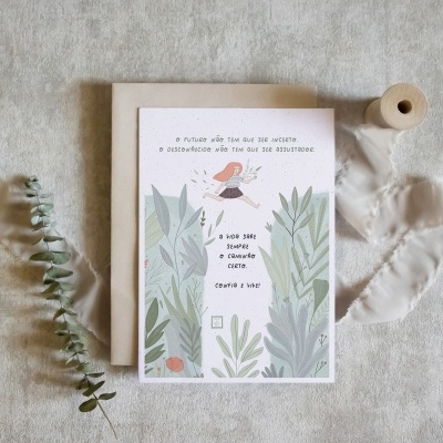 Cartão ilustrado com personagem e folhas verdes sobre tecido bege com envelope, ramo de folhas e fita enrolada.