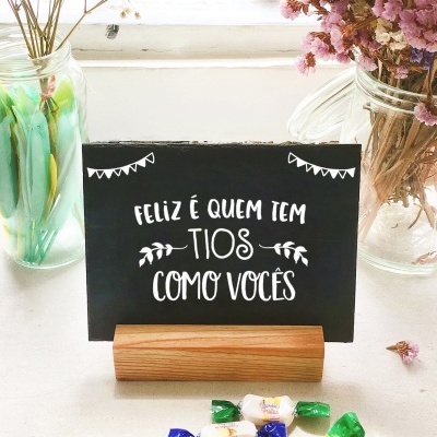 Cartão preto com mensagem para tios, vasos com flores e doces sobre mesa branca