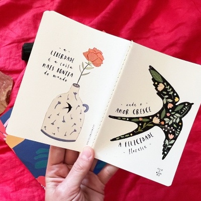 Caderno aberto com ilustrações de vaso com flor e pássaro, texto em português, fundo vermelho e pedra rosa.