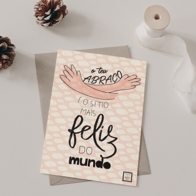 Cartão de felicitações com mensagem e envelope sobre mesa branca, acompanhado por pinhas e fita