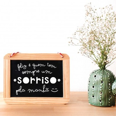 Quadro de madeira com texto em ardósia preta ao lado de vaso verde em forma de cacto com flores brancas