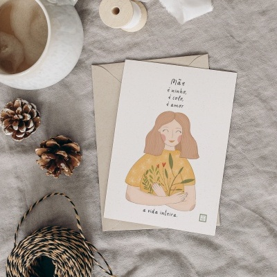Cartão com ilustração e texto sobre envelope, ao lado pinhas, rolo de linha e copo branco