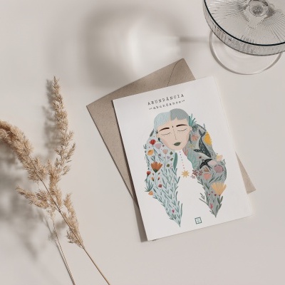Cartão ilustrado com mulher e flores, envelope castanho e raminhos secos numa superfície branca