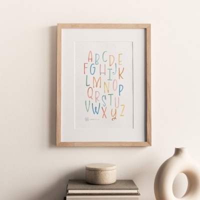 Poster de alfabeto colorido em moldura de madeira clara na parede, sobre mesa com caixa de cortiça, livros e vaso decorativo branco.