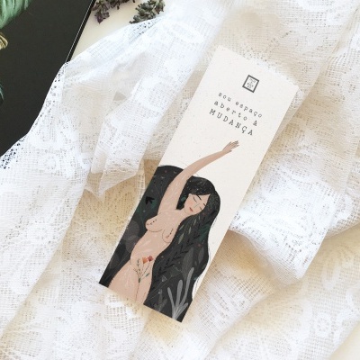 Caixa branca de produto com ilustração de mulher e texto, sobre tecido branco rendado junto a livro preto