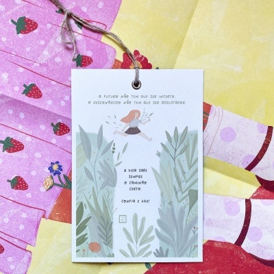 Cartão com texto em português pendurado sobre tecidos coloridos com estampas diversas.