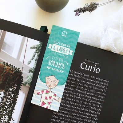 Marcador de livro com ilustração colorida e texto em português junto a livro aberto e planta suculenta