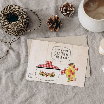 Cartão postal com ilustrações de utensílios de cozinha e texto motivacional sobre tecido cinza, junto a objetos de costura e café.