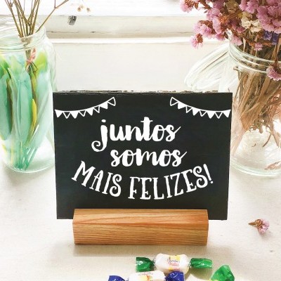 Placa de madeira preta com texto em branco entre dois vasos de vidro com flores secas e balas coloridas numa mesa branca