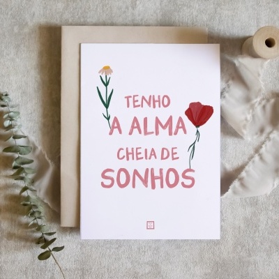 Cartão/postal branco com texto em vermelho e flores ilustradas, colocado sobre papel castanho com fita branca e ramo verde.